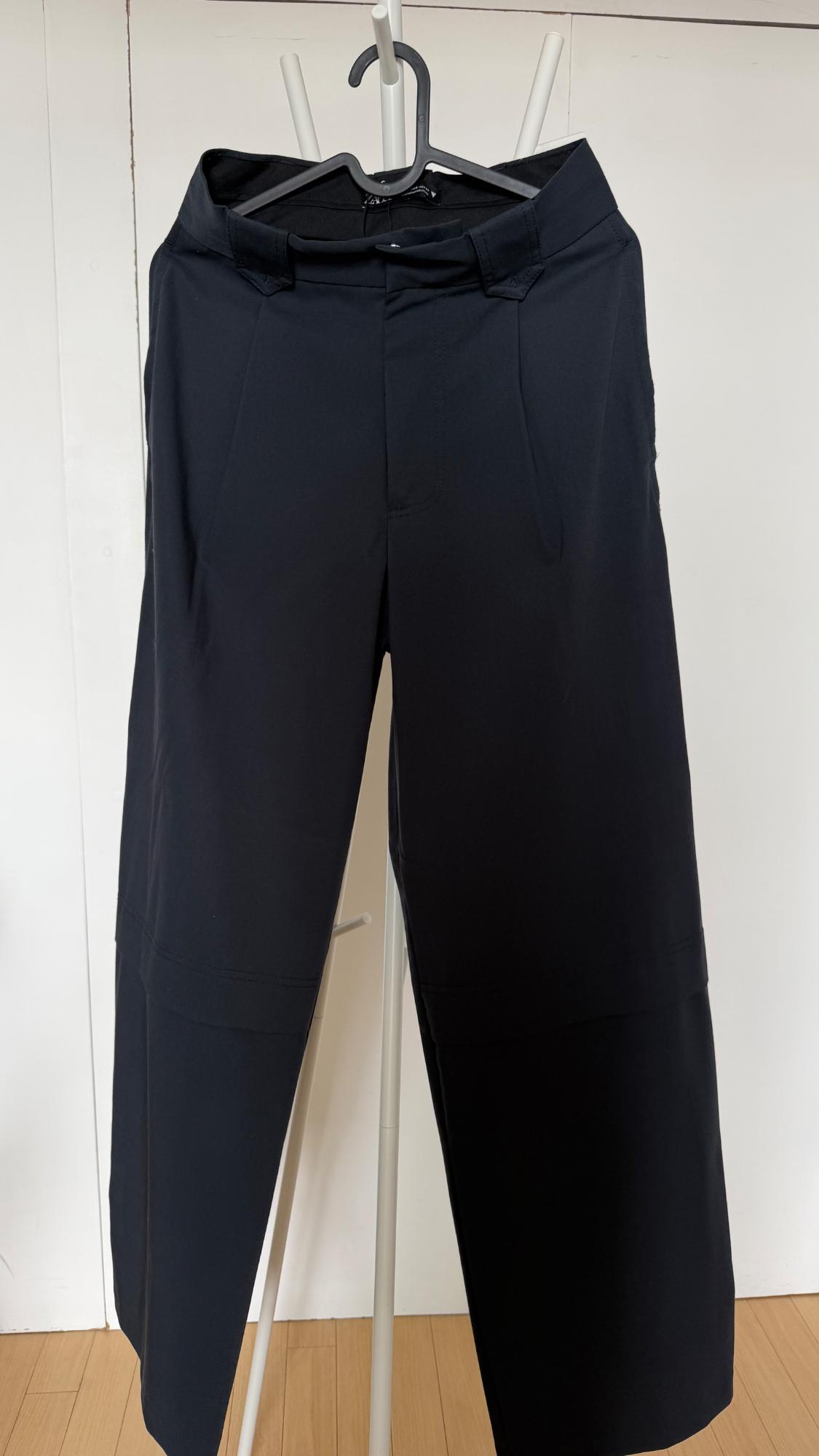 Black trousers with a horizontal seam on the leg, hanging on a hanger on a white clothes rack against a background of white doors.