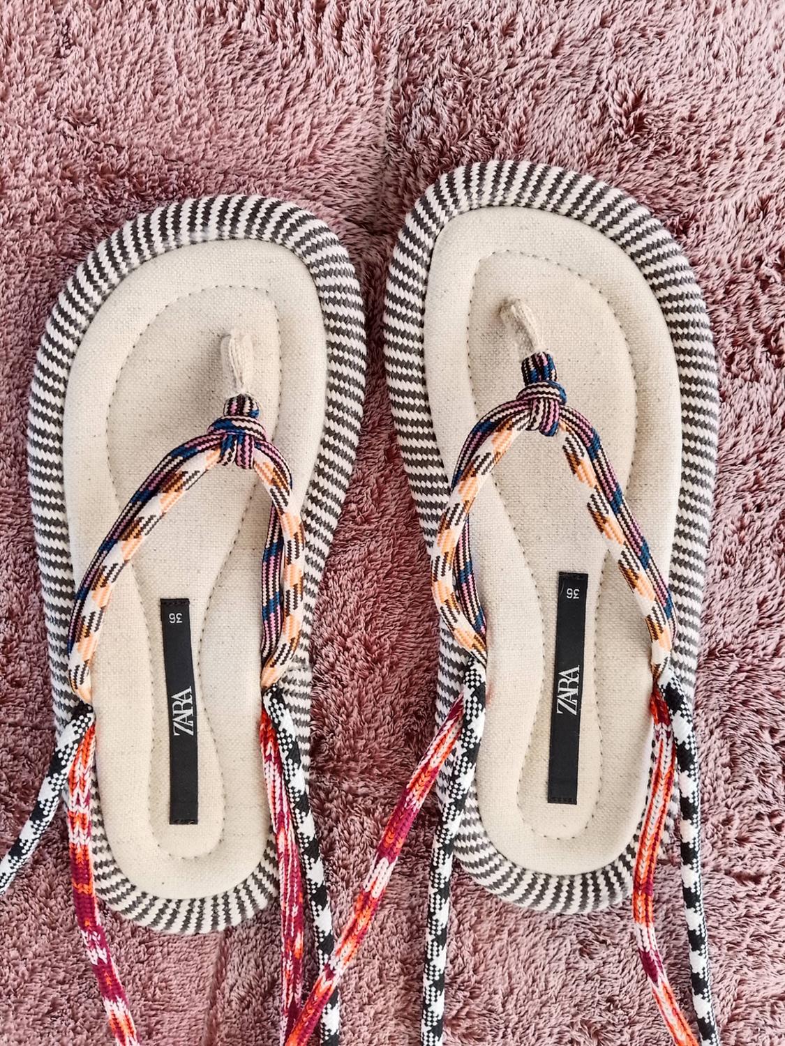 Sandals with multi-coloured braided straps and a striped trim on a soft-textured pink background. Text: ZARA 36.