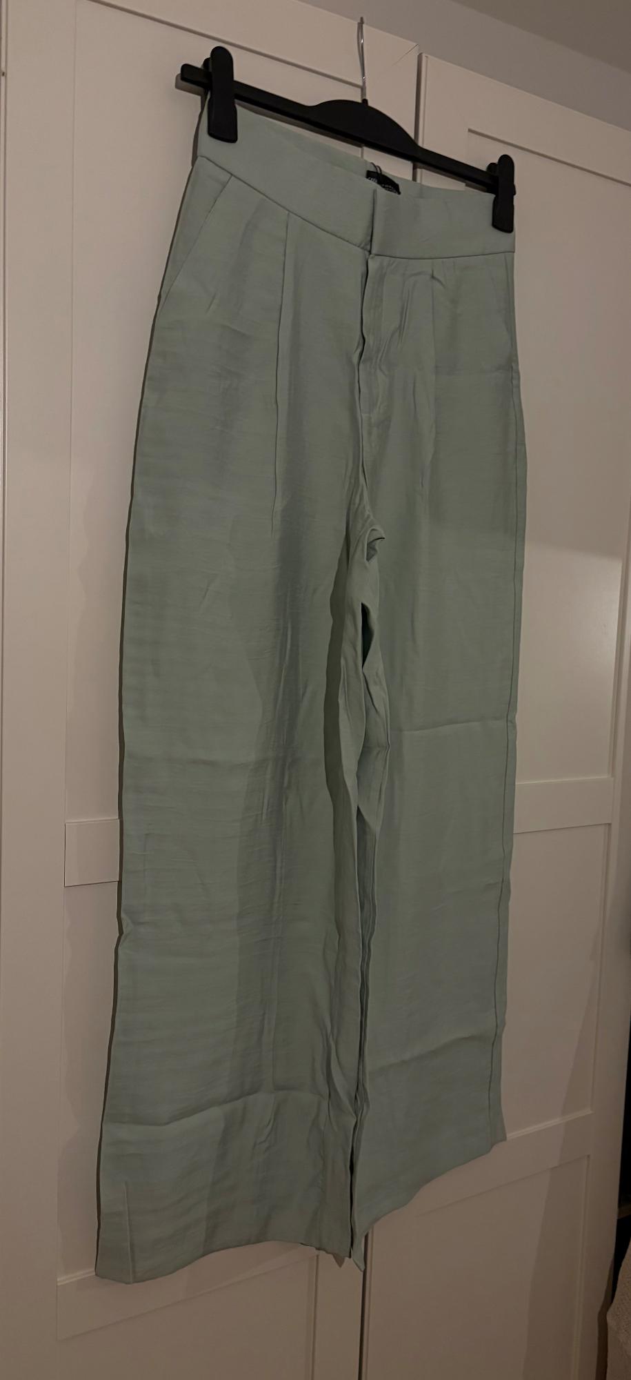 Light green long trousers hanging on a black hanger against a white wardrobe background.