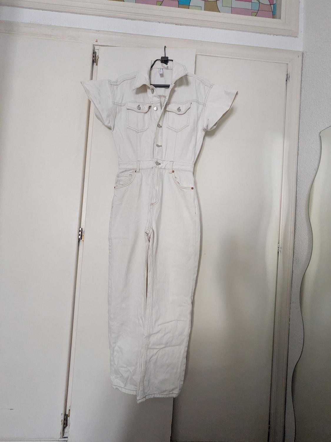 White short-sleeved denim jumpsuit with buttons and pockets, hanging on a hanger in front of a white wardrobe in a relaxed setting.