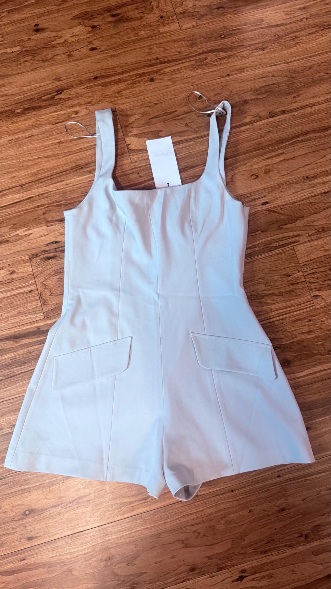 Light blue short jumpsuit with wide straps, flap pockets, and front seams, laid out on a wooden floor.