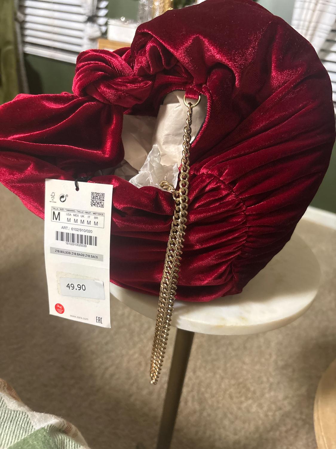 Red velvet purse with gold chain on white table. Visible tag with price of 49.90. Background with blinds and furniture.