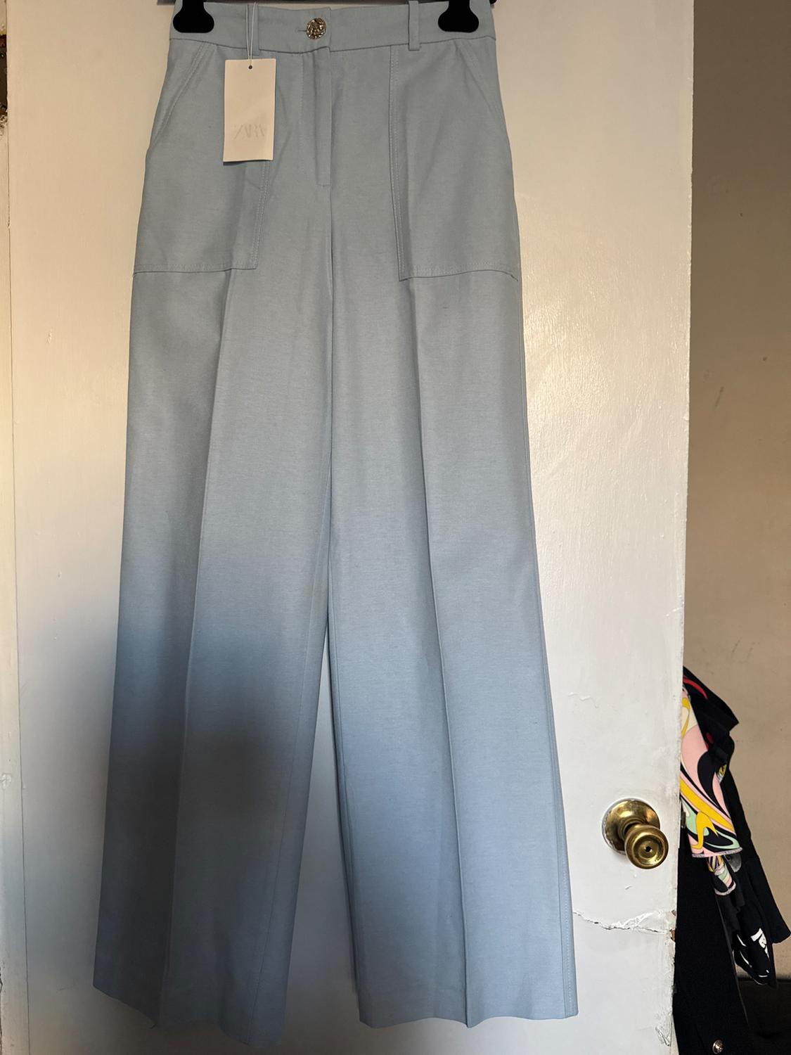 Light blue pants hanging on a white door, with a visible tag. Background with a gold doorknob and partially visible colorful clothing.