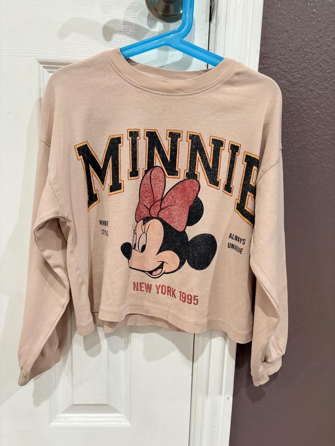 Beige sweatshirt with Minnie Mouse print, text "MINNIE", "NEW YORK 1995", "MINNIE STYLE", "ALWAYS UNIQUE", hanging on a blue hanger.