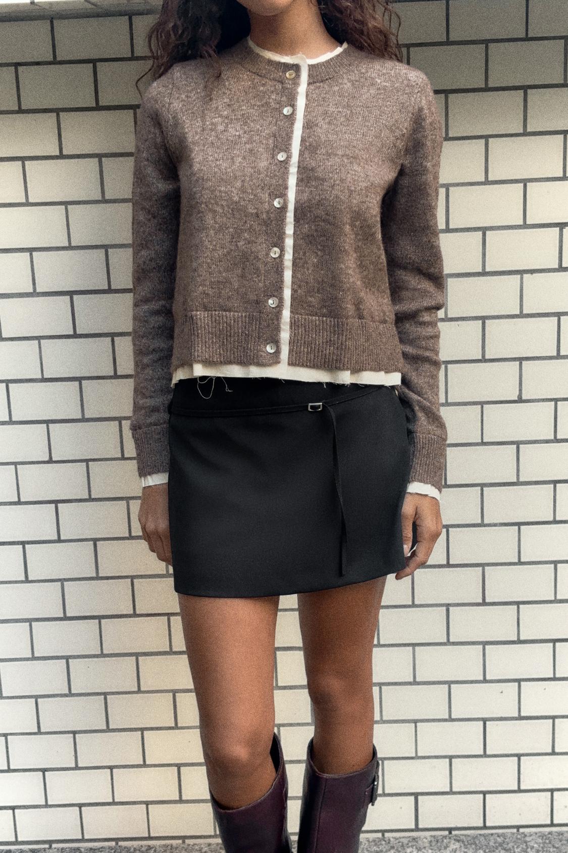 Komplet rjavega jopice na gumbe in kratkega črnega krila, kombiniran z rjavimi visokimi škornji.