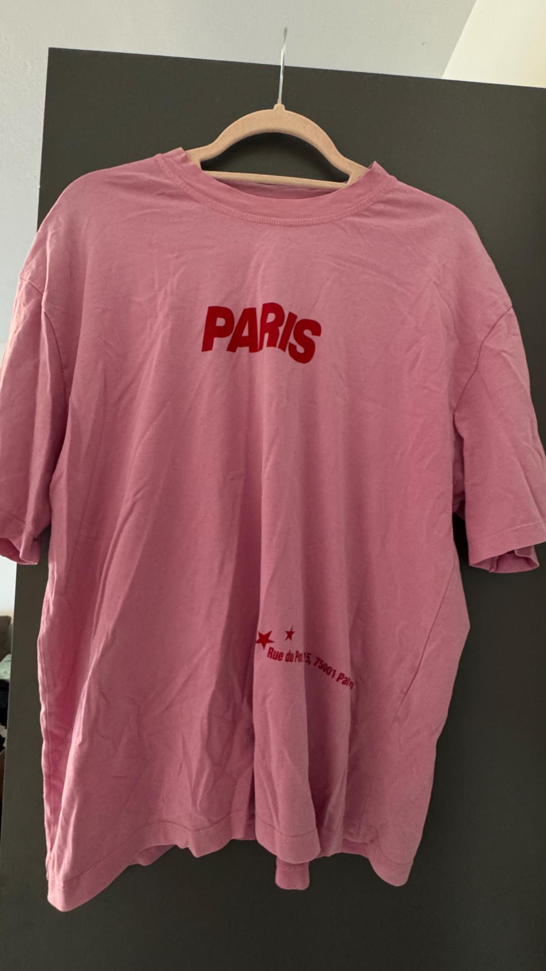 Pink t-shirt hanging on a hanger with the word "PARIS" in red and additional text at the bottom. Dark background.