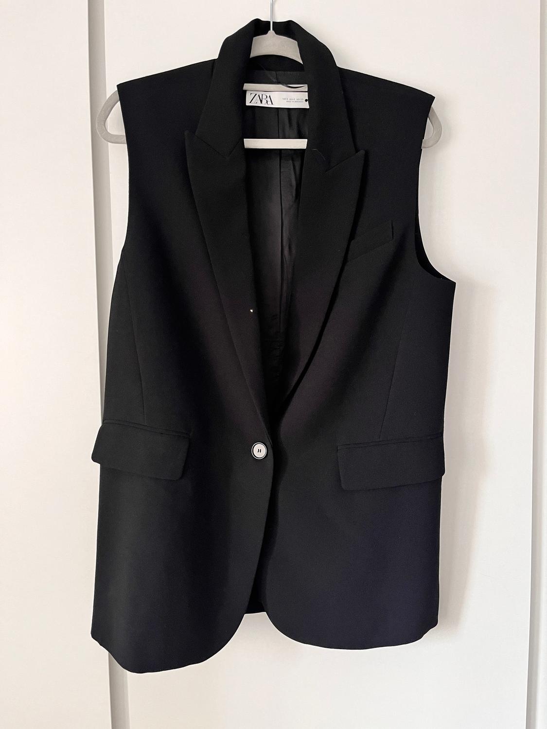 Black sleeveless waistcoat hanging on a white hanger, with lapels and one button, visible label with the text "ZARA".