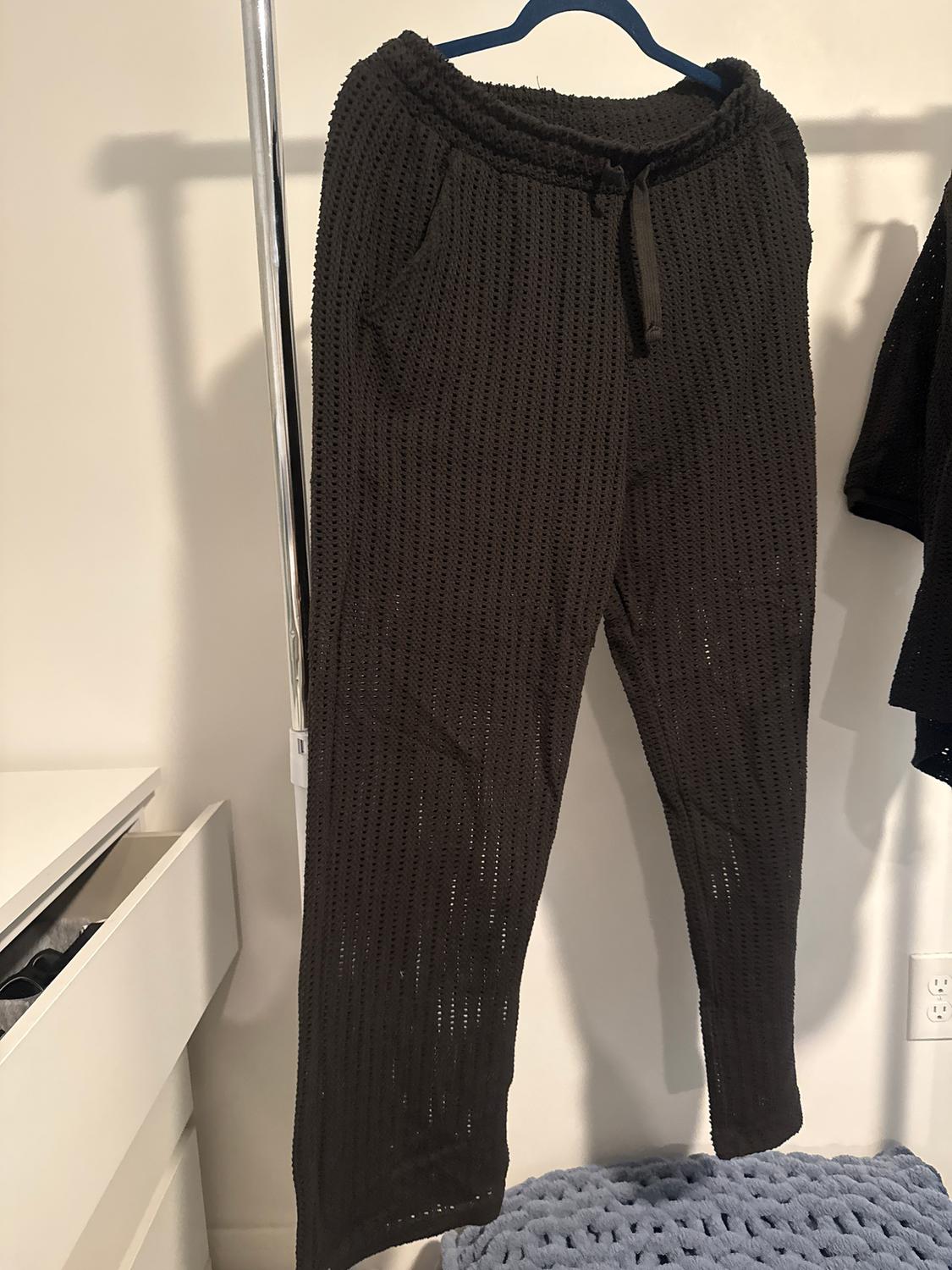 Dark-coloured open-knit trousers hanging on a blue hanger. Background with a white wall and an open chest of drawers.