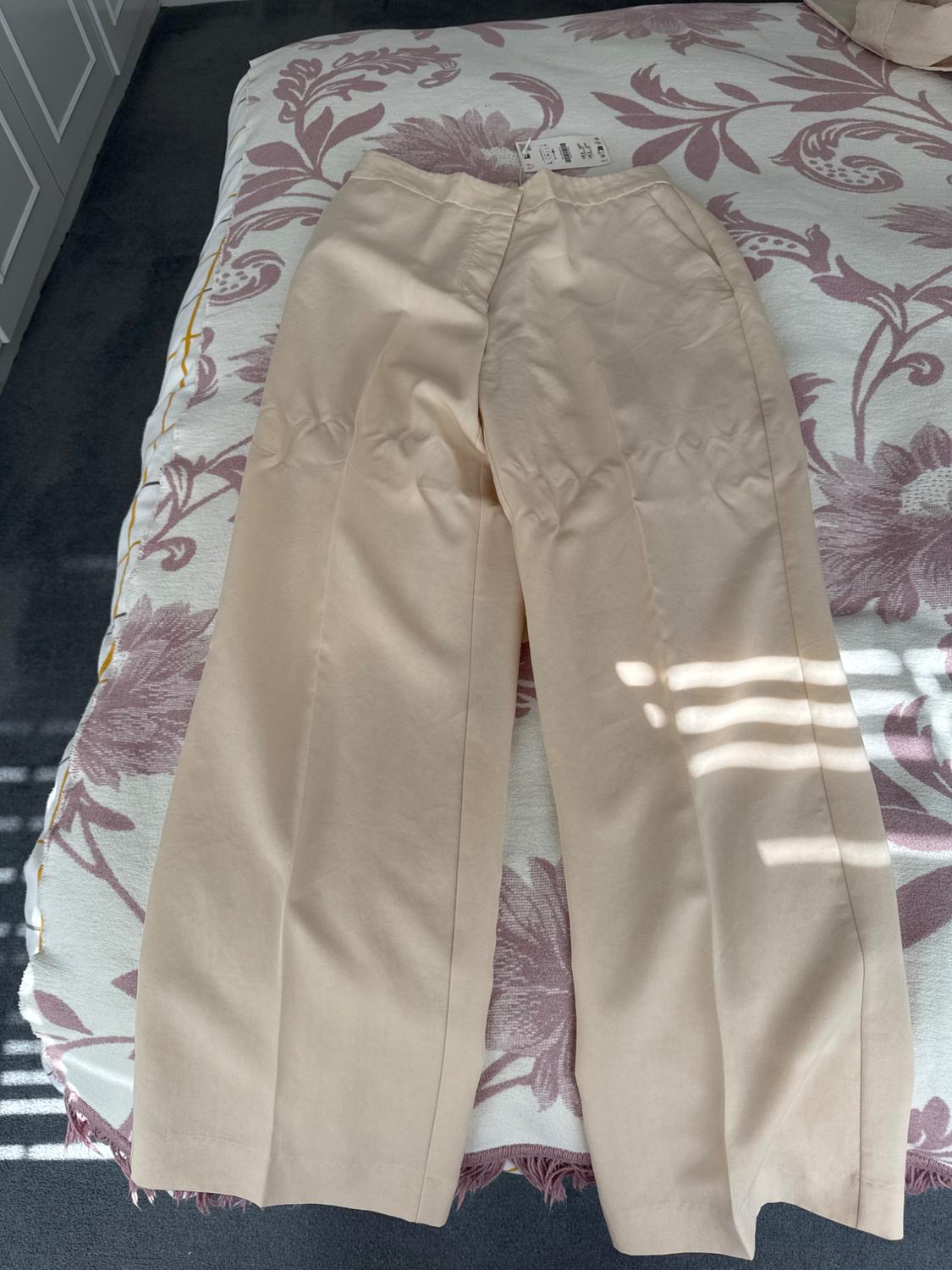 Beige trousers on a bed with a purple floral bedspread. Label visible on the waistband. Natural light coming through the window.