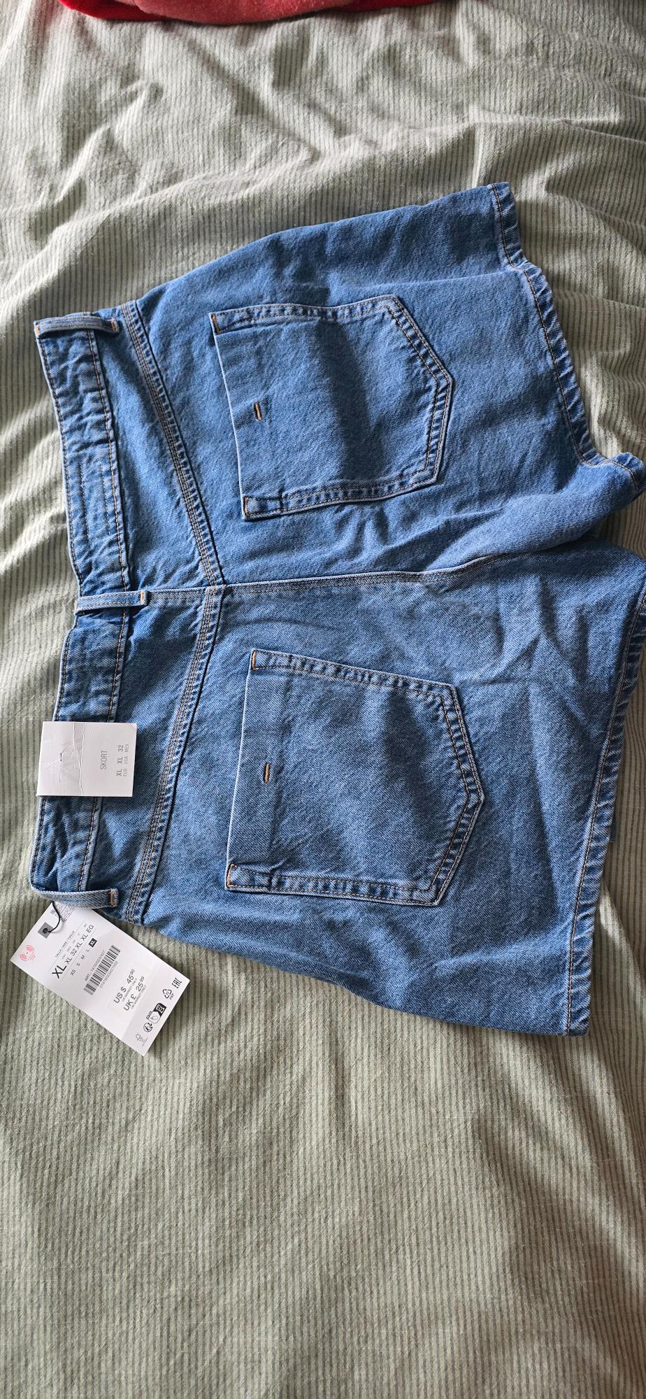 Blue denim shorts with two back pockets, hang tags visible on a striped quilt.