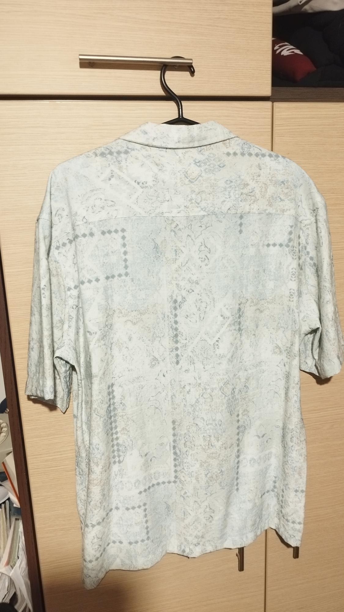 Short-sleeved shirt with a subtle light-toned print, hanging on a hanger in front of a light wood wardrobe.