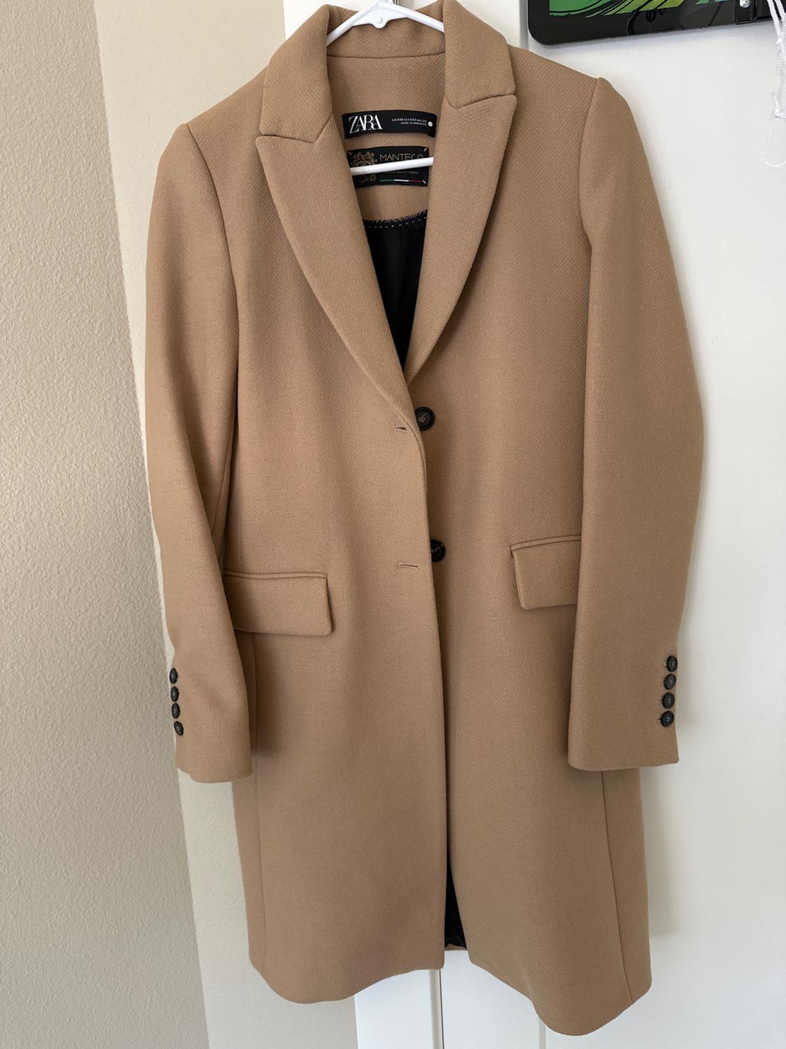 Camel coat hanging on a white hanger, with lapels and black buttons. Visible Zara label on the inside of the neck.