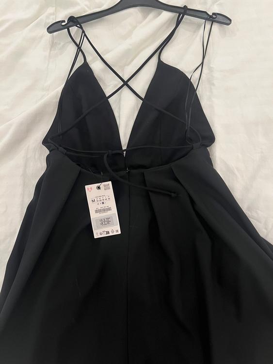 Black dress with cross-back straps, hanging on a black hanger against a white fabric background. Visible tag with price and size.