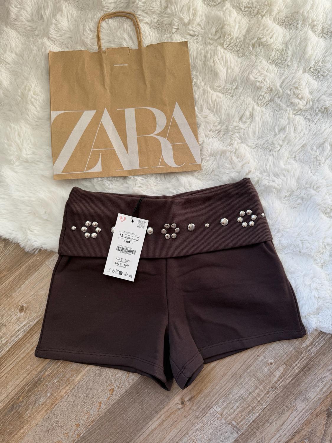 Brown shorts with studs and a tag, beside a paper bag bearing the text ZARA, on a white rug and wooden floor.