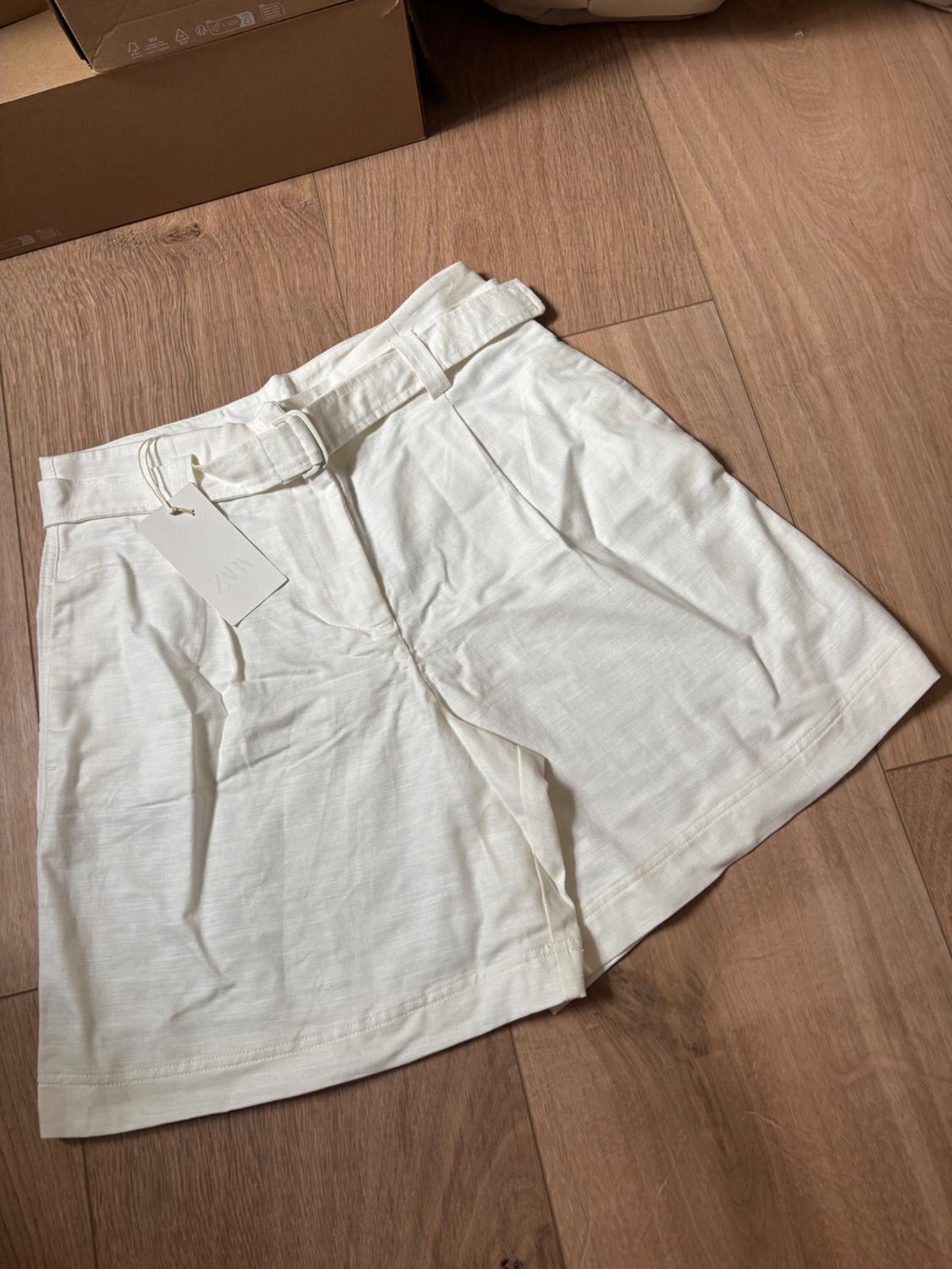 White shorts with a belt on a wooden floor. Label with the text ZARA. Casual style with cardboard boxes in the background.