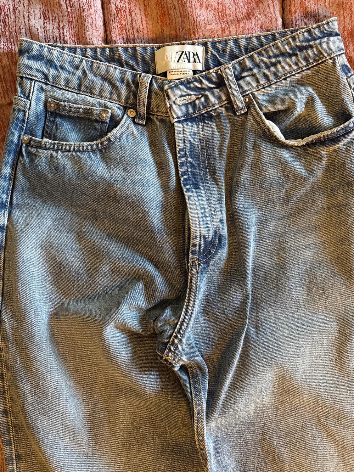 Close-up of blue jeans on a reddish fabric. The waistband, pockets, and inner label with the text ZARA are prominent.