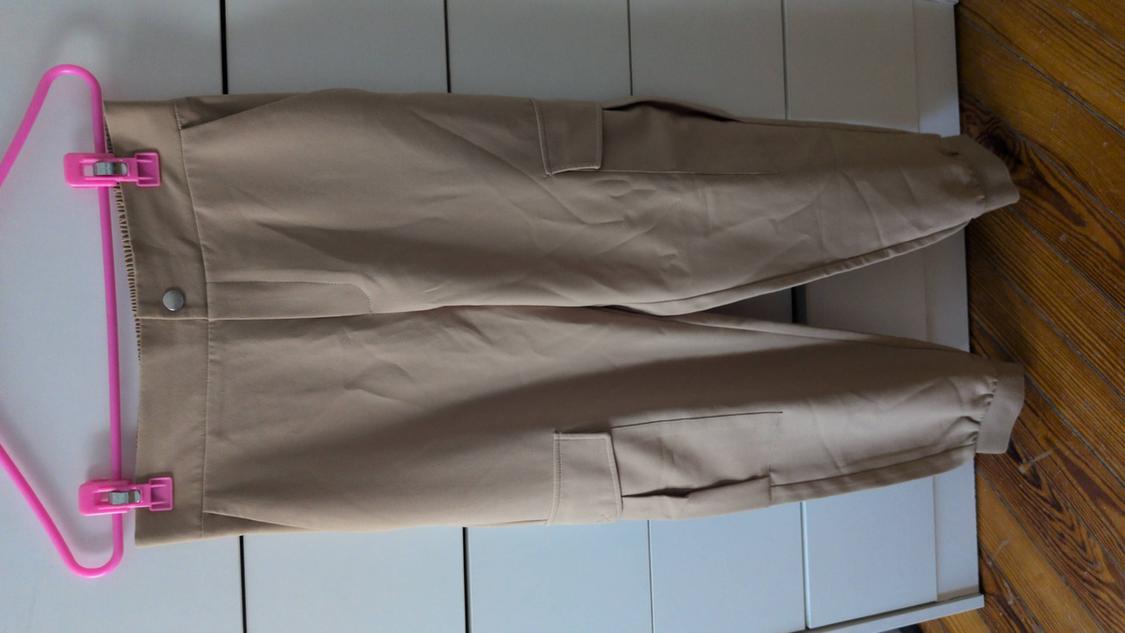 Beige cargo trousers hung on a pink clothes hanger with clips, against a background of white drawers and a wooden floor.