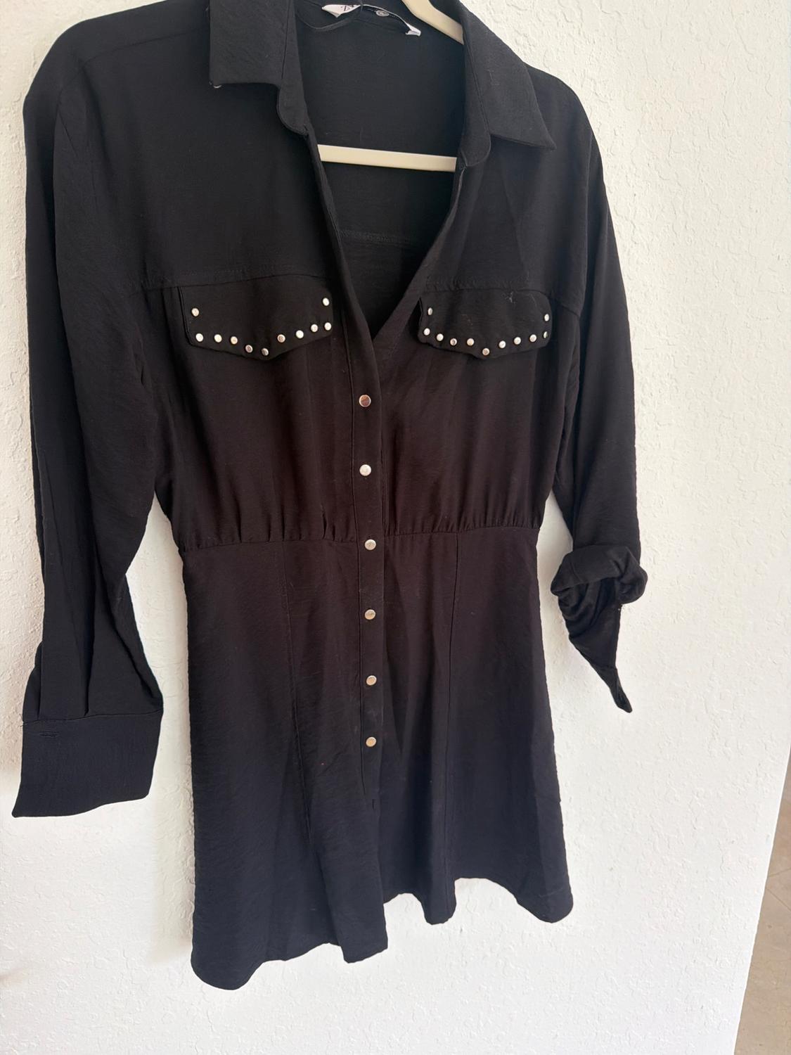 Black long-sleeved shirt dress on a hanger against a white wall. Casual style with a fitted waist, buttons and studs on the pockets.