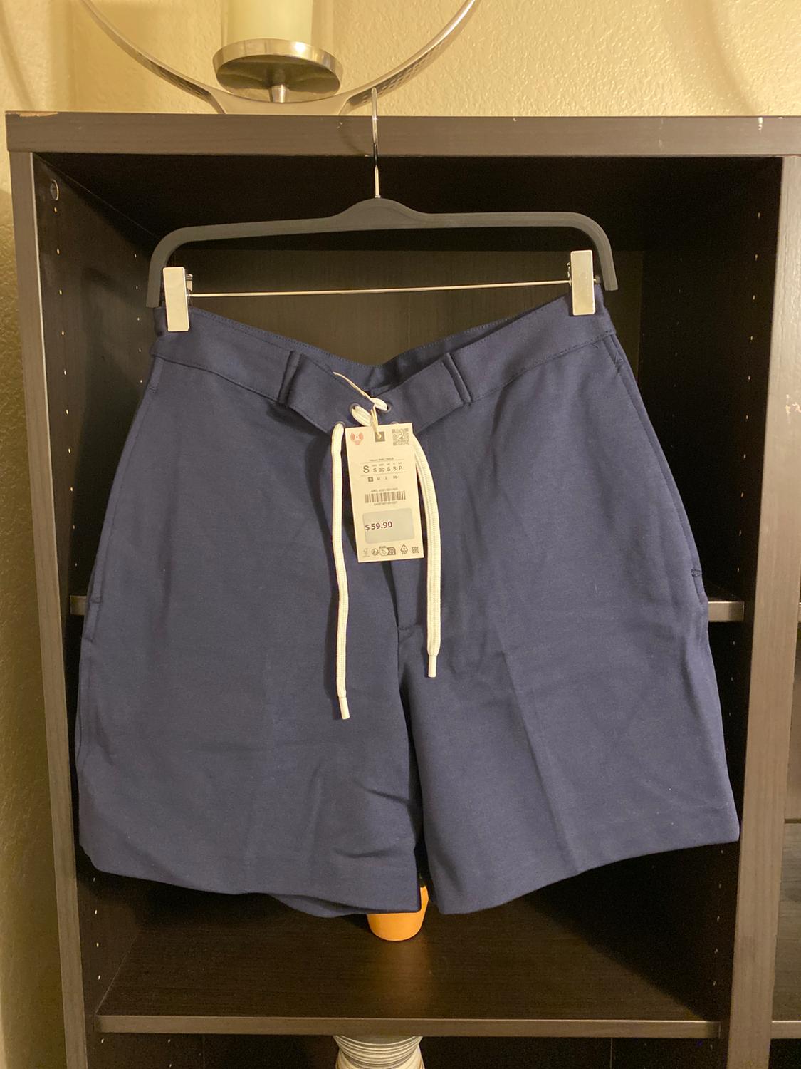 Navy blue shorts with a white drawstring, hanging on a black hanger. Visible tag with a price of $59.90. Shelf background.