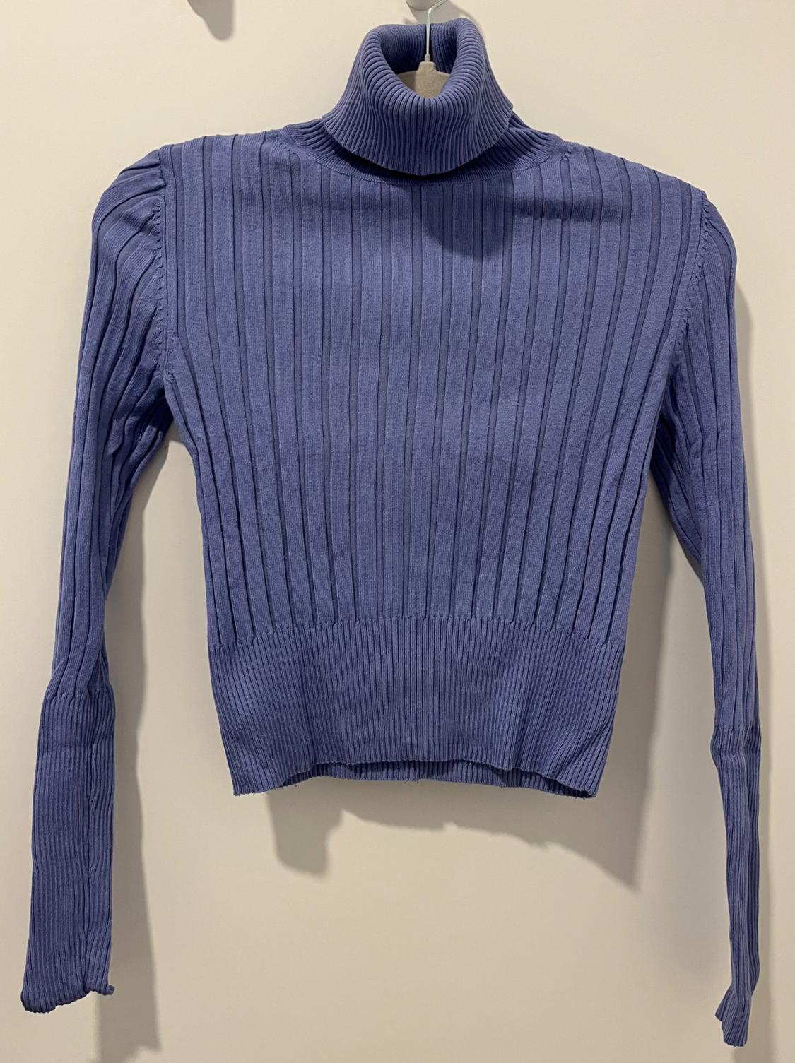 Blue knitted jumper with a high neck and long sleeves, hanging on a hanger against a beige background.