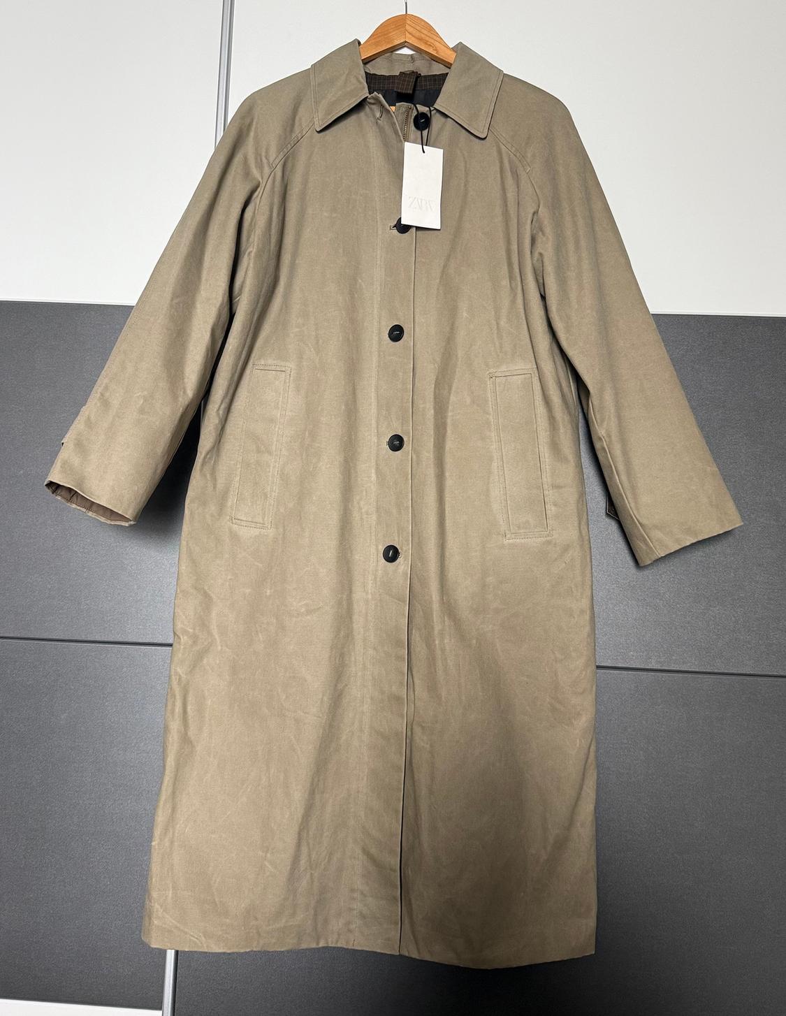 Long beige coat with black buttons hanging on a wooden hanger. Visible label with the text "ZARA". Grey and white background.