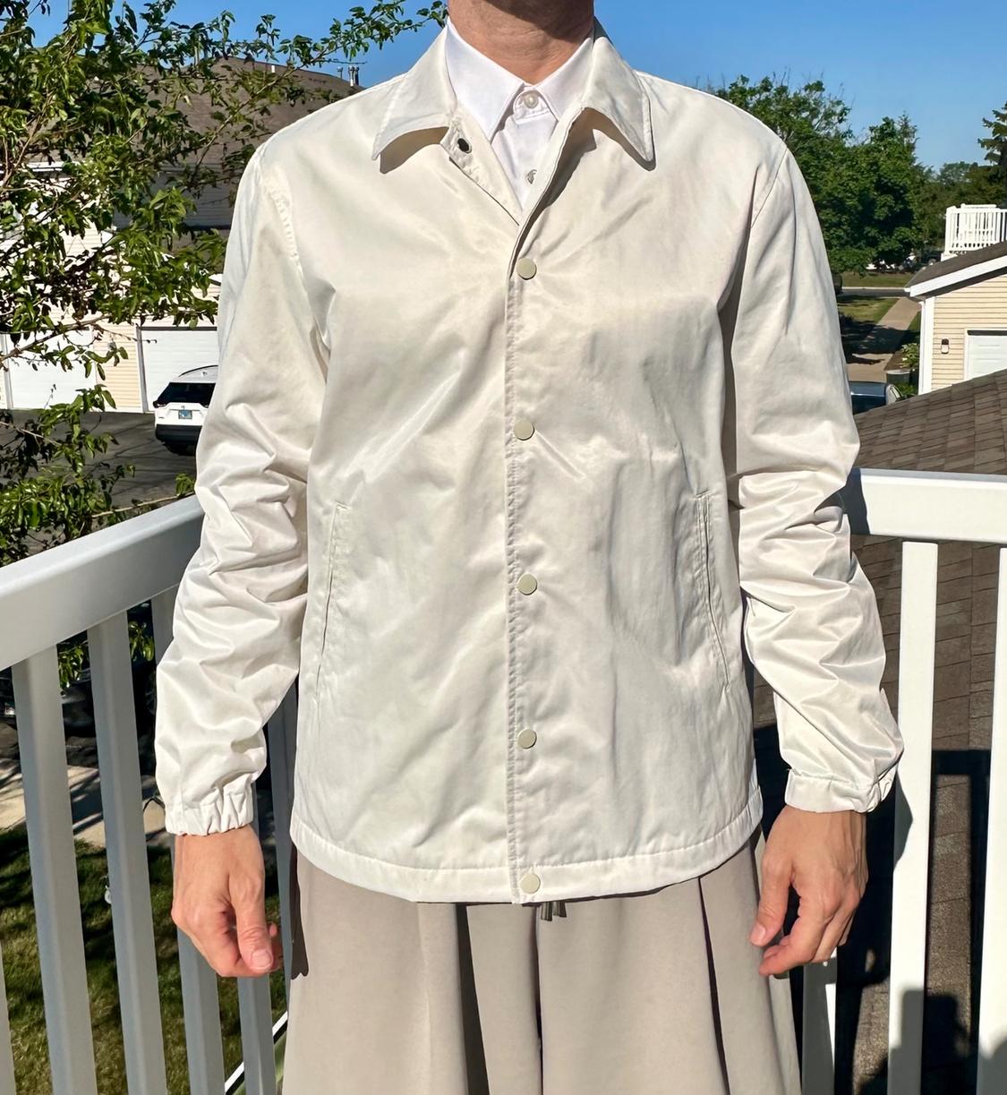 Man in a white button-up jacket and white shirt, standing on an outdoor balcony with trees and houses in the background.