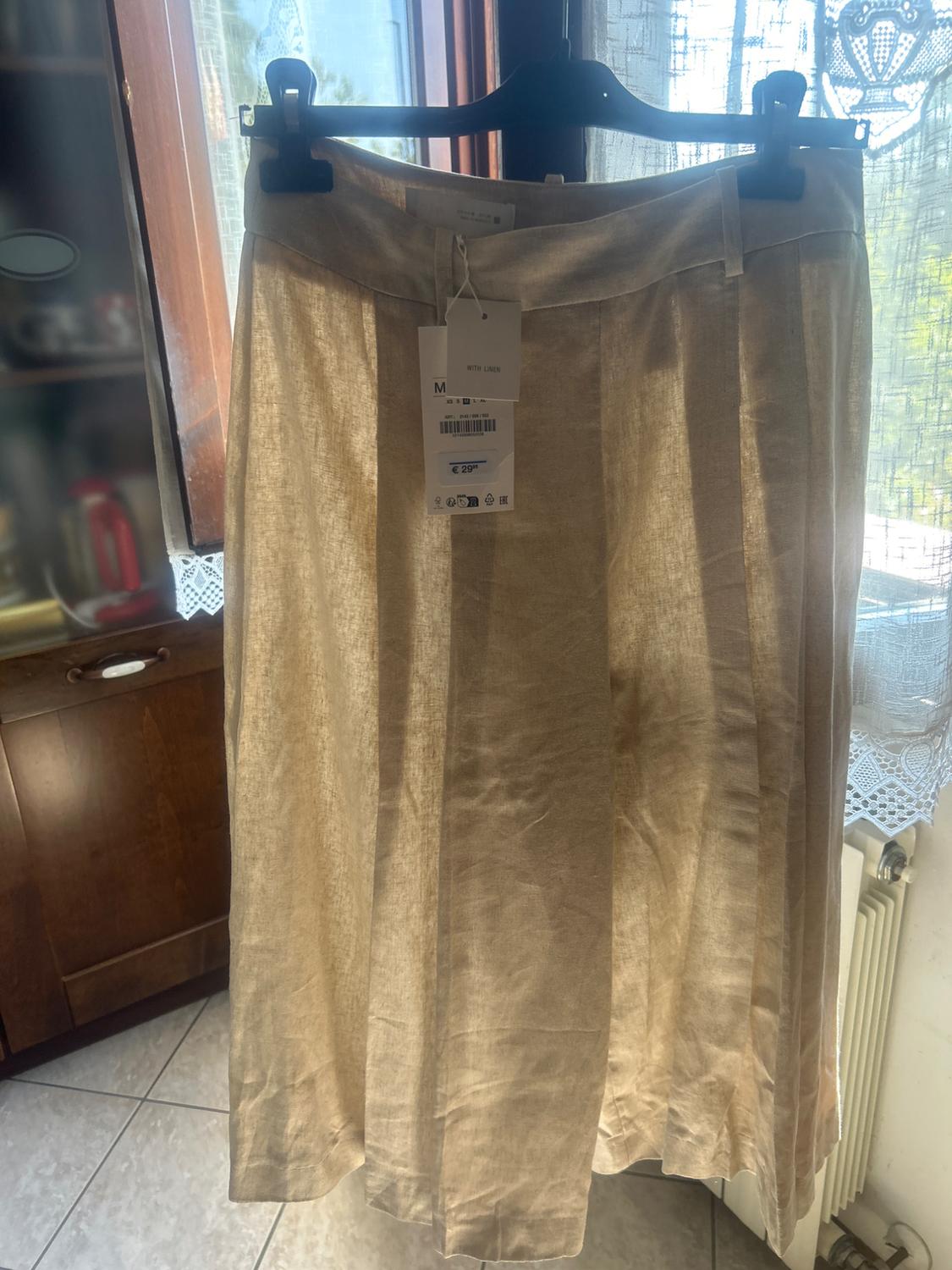 Beige skirt hanging on a hanger in front of a window. Visible label with text: "WITH LINEN". Kitchen background with natural light.