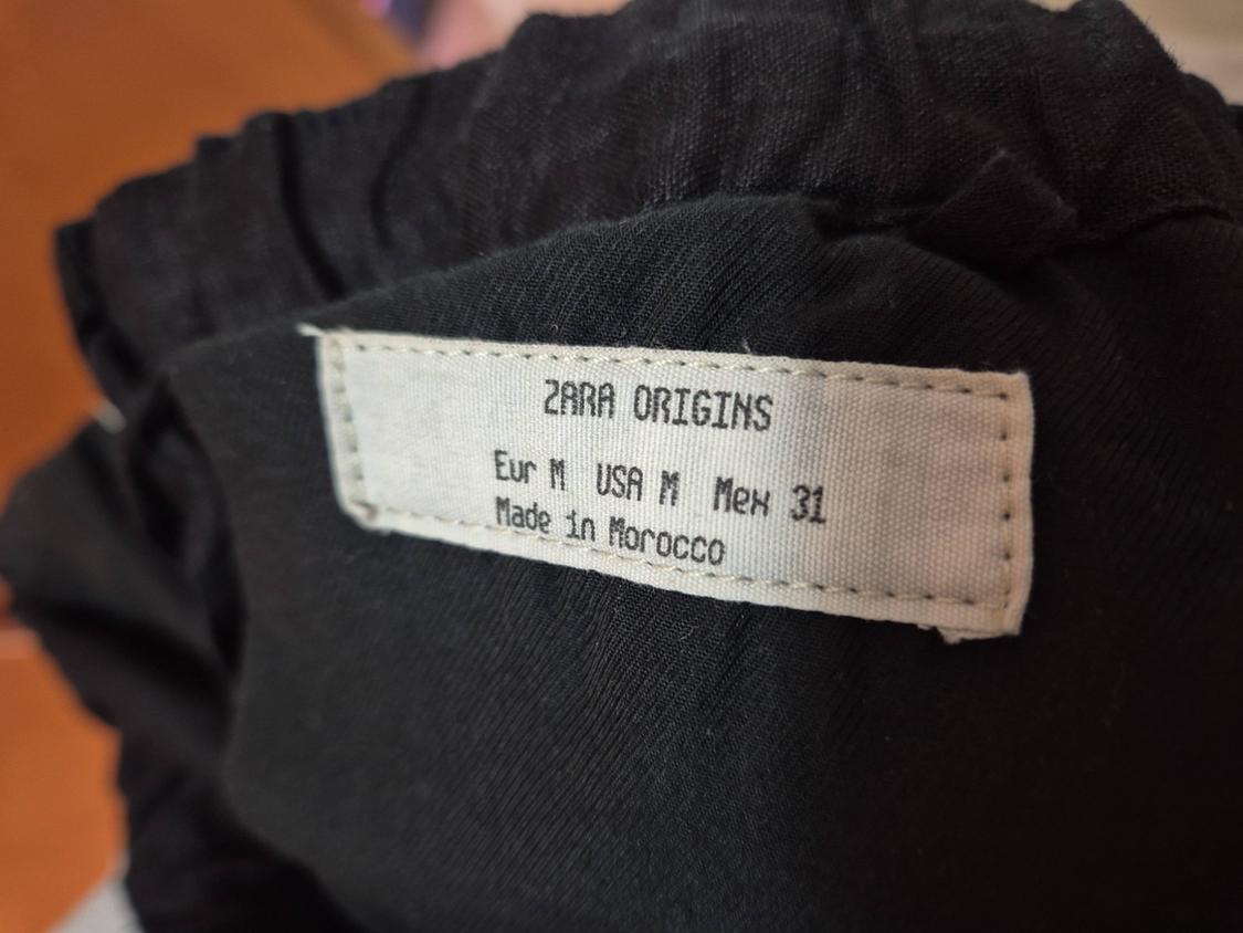 Black clothing label with text: "ZARA ORIGINS, Eur M USA M Mex 31, Made in Morocco". Blurred background.