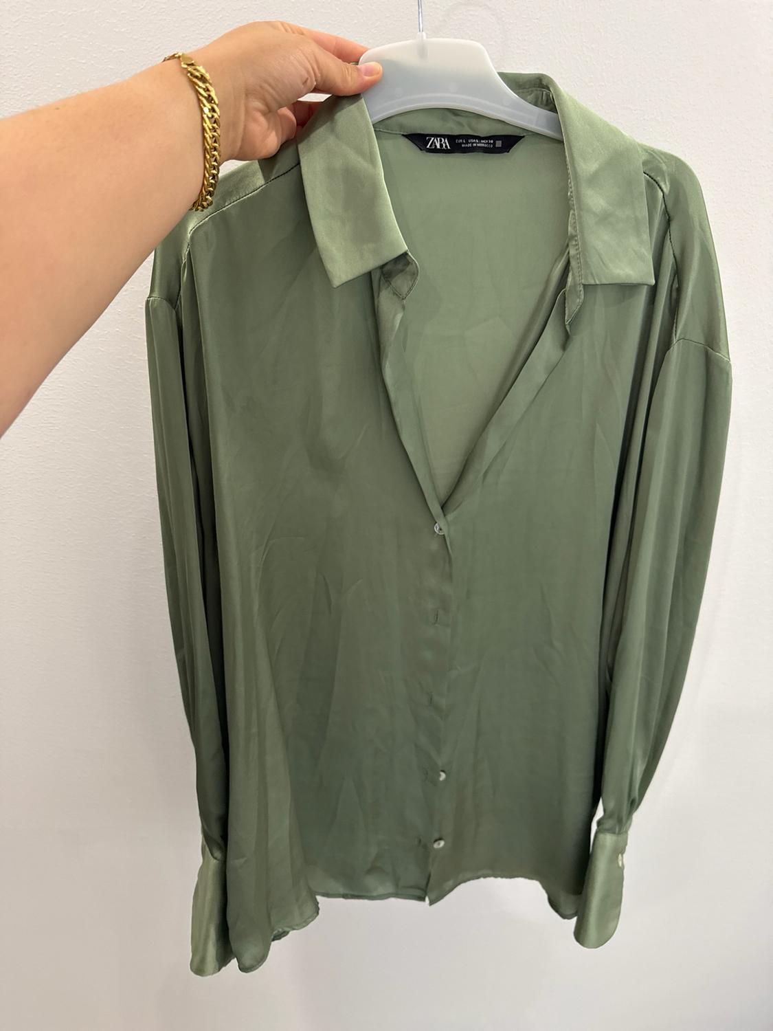 Hand with a gold bracelet holding a hanger with a green satin long-sleeved shirt. Label with the text ZARA. White background.
