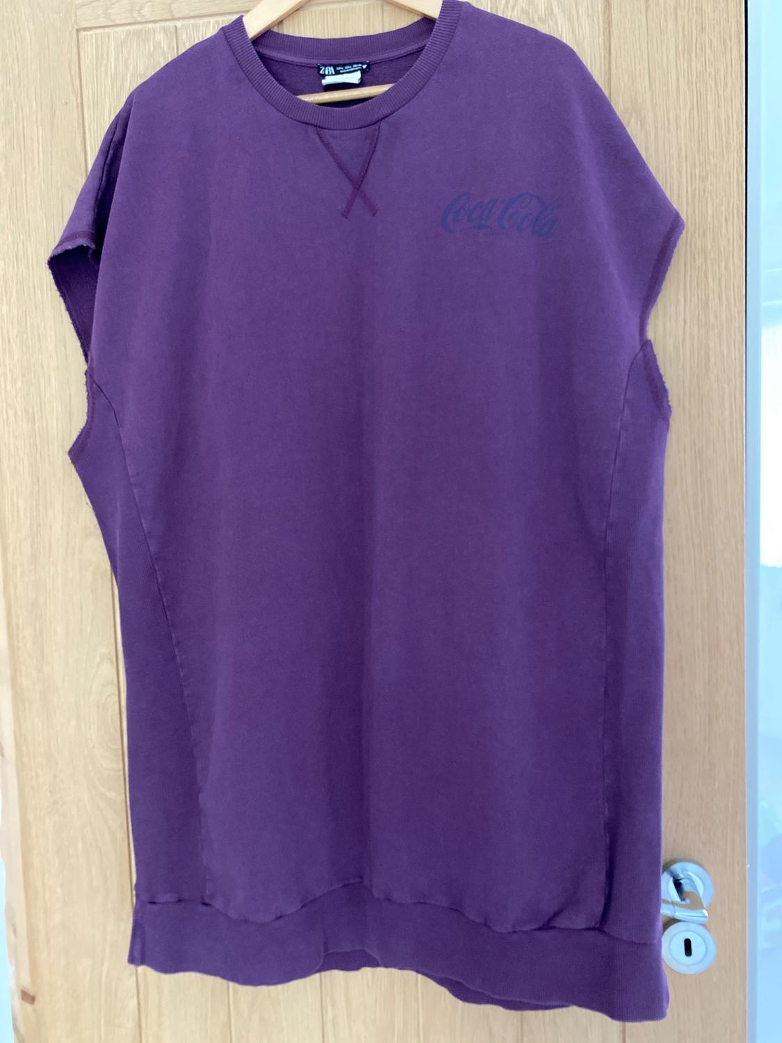 Purple sleeveless T-shirt hanging on a wooden door. It has the Coca-Cola logo on the chest.