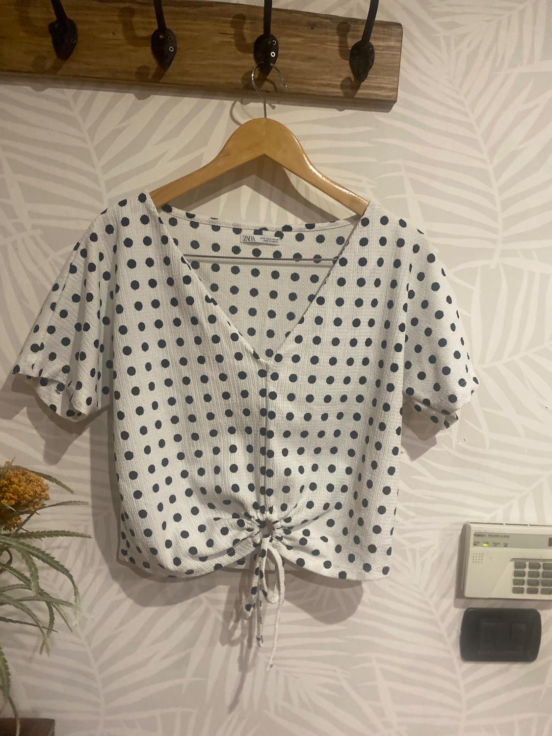 Front view of a white blouse with black polka dots, a V-neck and an adjustable knot at the bottom.