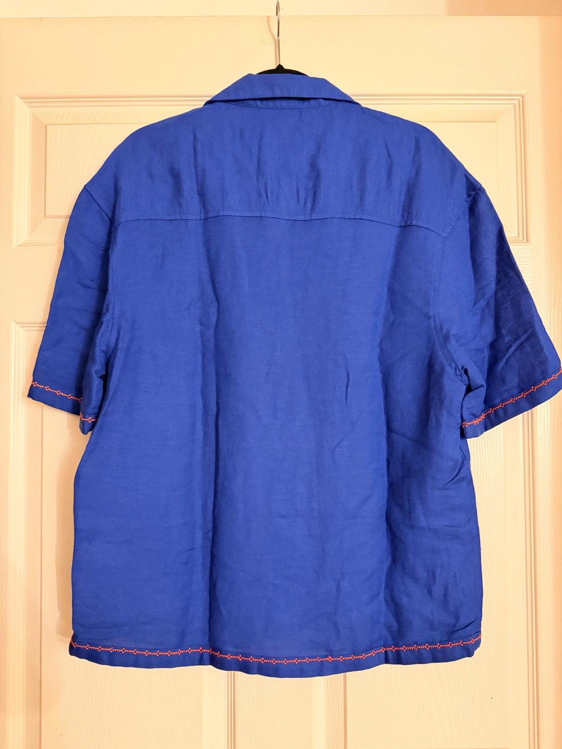 Blue short-sleeved shirt viewed from the back, hanging on a light-colored door. Red embroidered details on the sleeves and hem stand out.