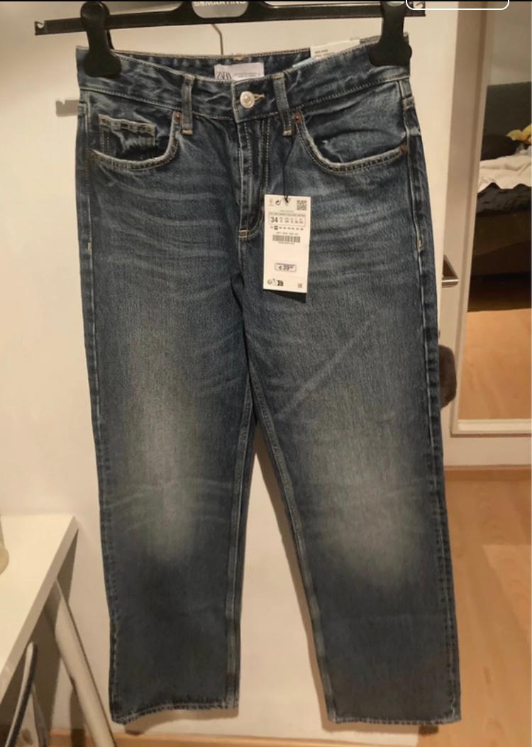 Blue jeans hanging on a black hanger, with a visible label in the centre. Room background with mirror and bed.