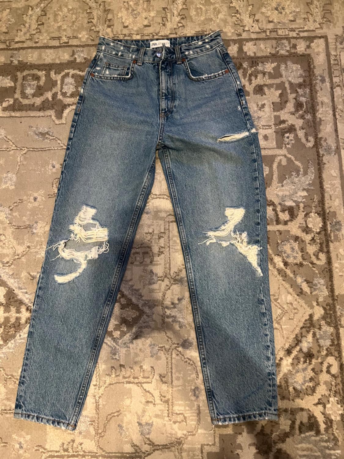 Distressed blue jeans with rips on the legs, placed on a geometric-patterned rug in beige and brown tones.