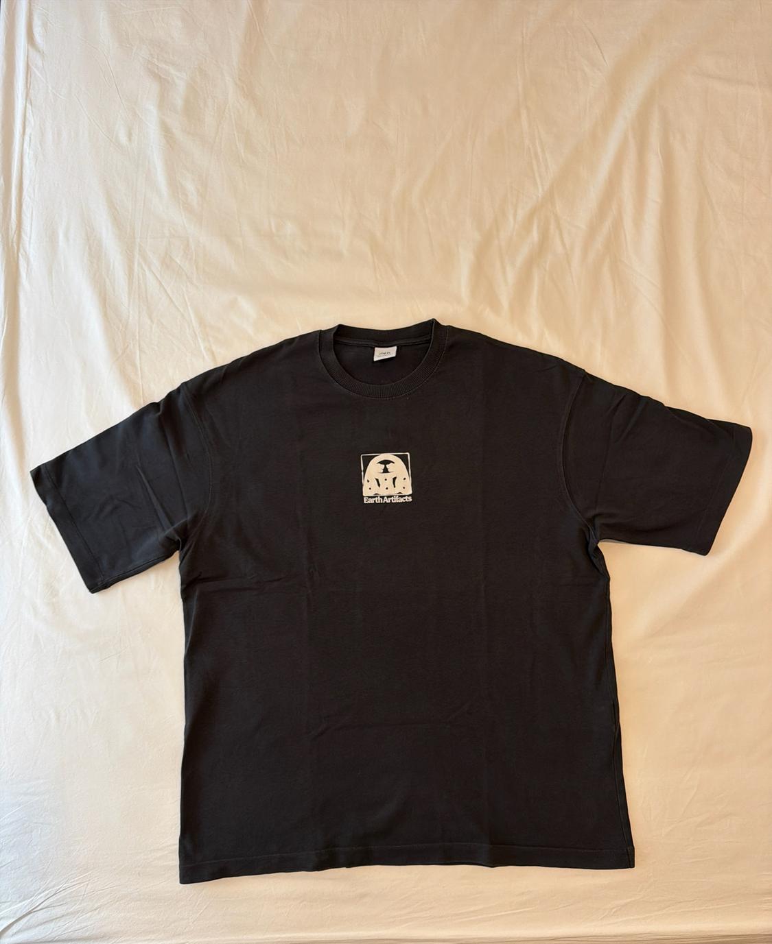 Black short-sleeved T-shirt on a crumpled white sheet. It has a central graphic with the text 'Earth Artifacts'.