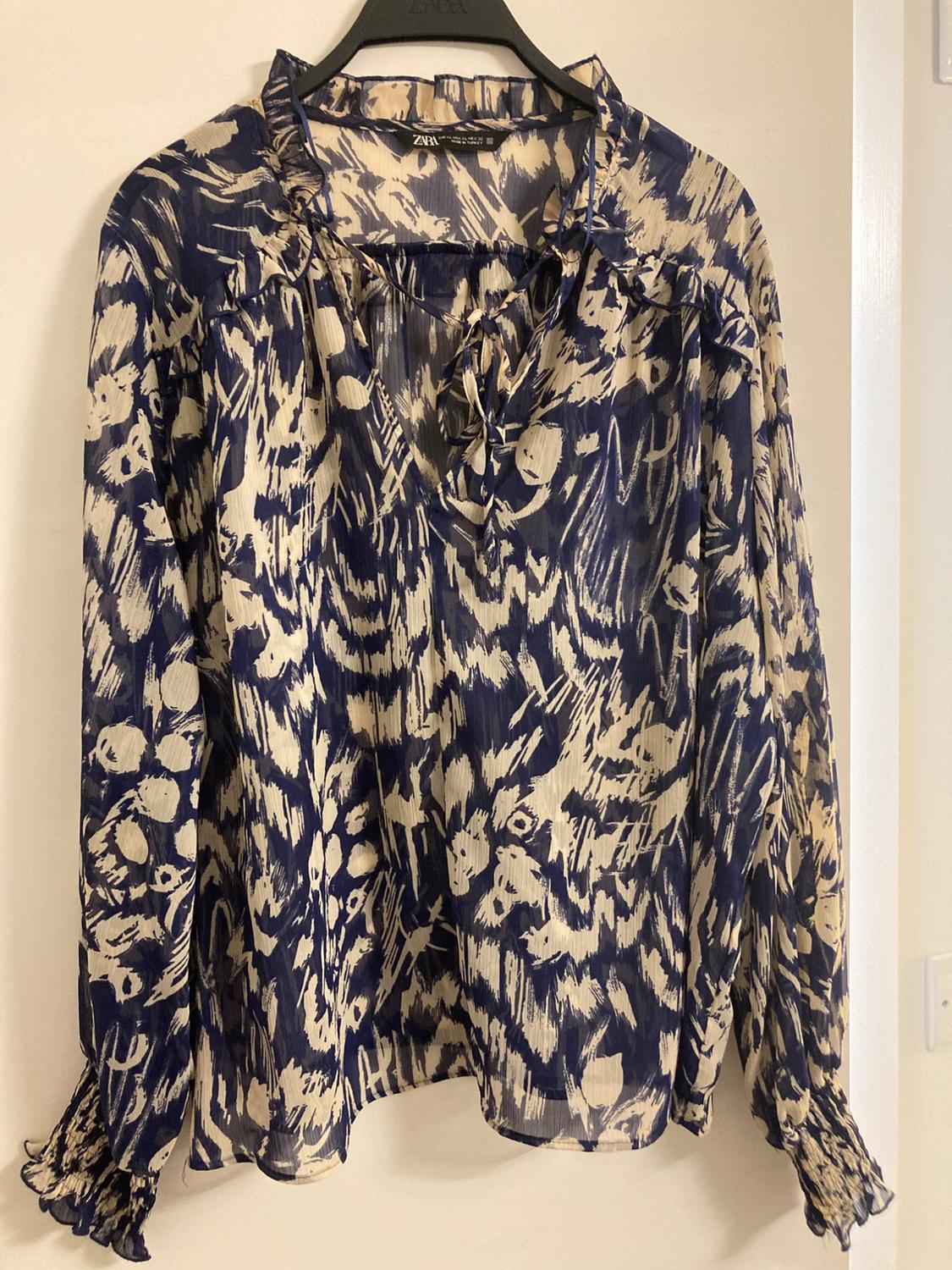 Blouse hanging on a black hanger, blue and beige abstract print, with neck tie and long sleeves with gathered cuffs.