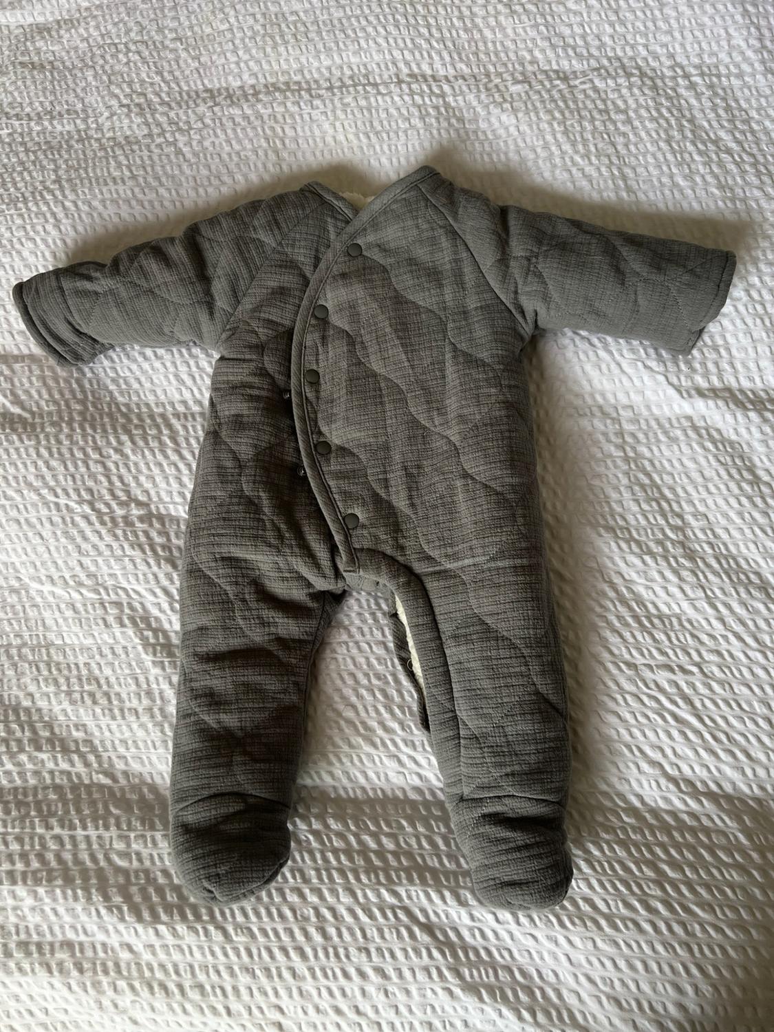 Grey padded baby snowsuit with enclosed feet and asymmetrical button fastening, on a white textured quilt. Cosy atmosphere.