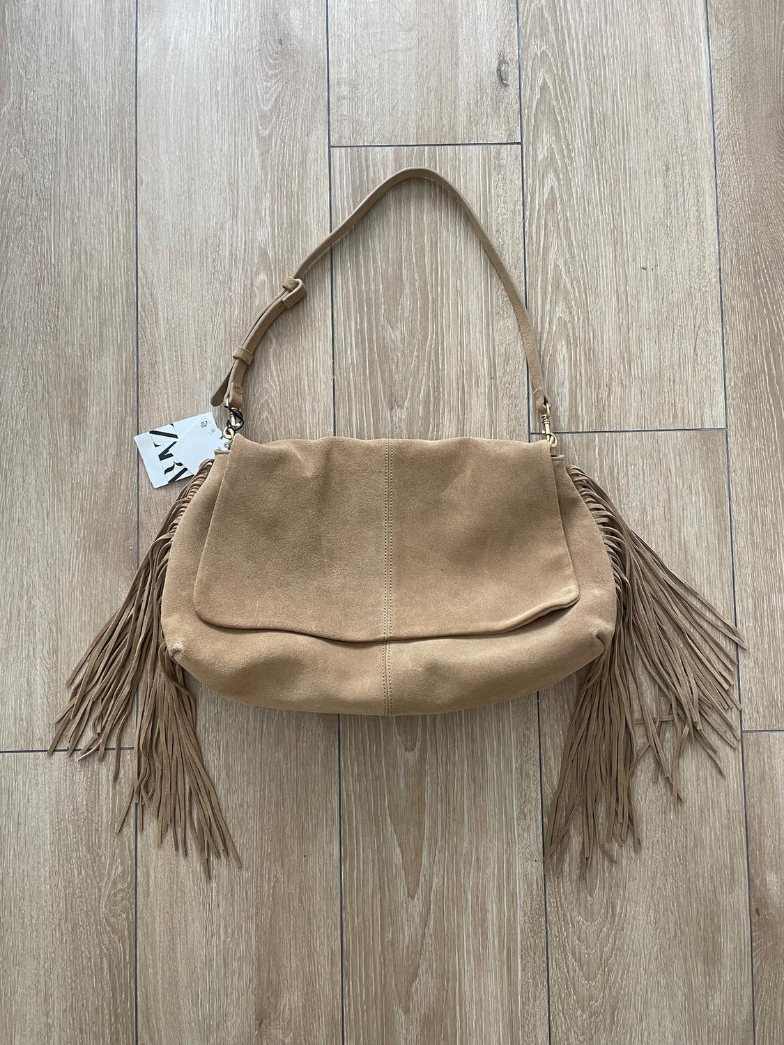 Beige handbag with fringes on the sides, on a light wooden floor. White tag with "Zara" text hanging from the adjustable handle.