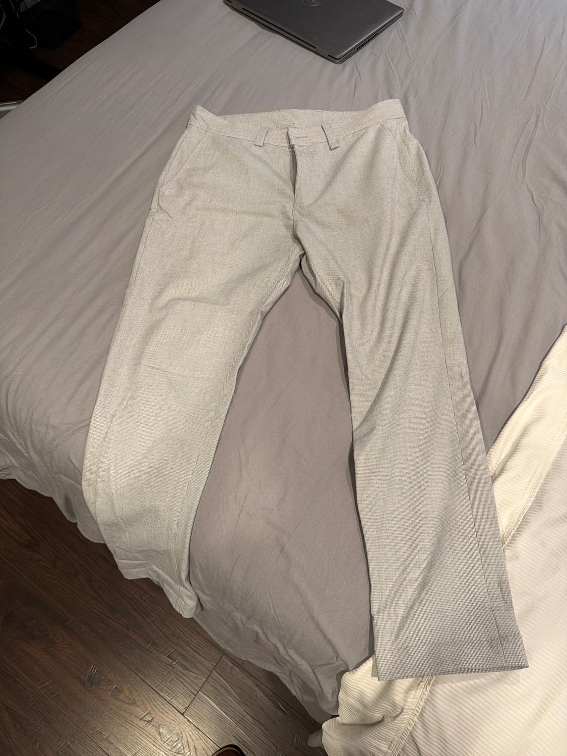 Light grey trousers laid out on a bed with grey sheets. Relaxed setting with a laptop and wooden floor visible.