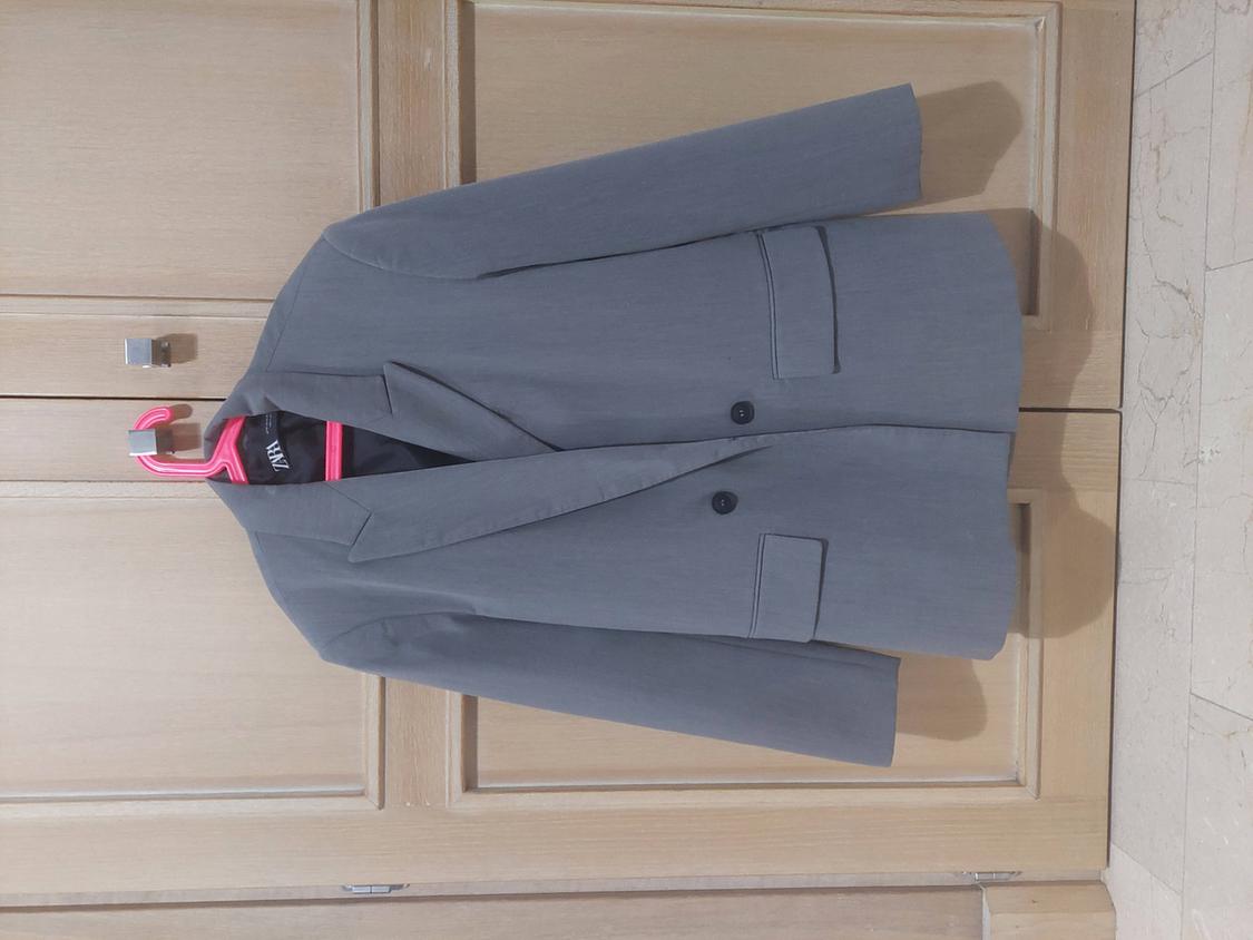 Grey jacket hanging on a pink hanger in front of a light wooden door. The inside label is visible.