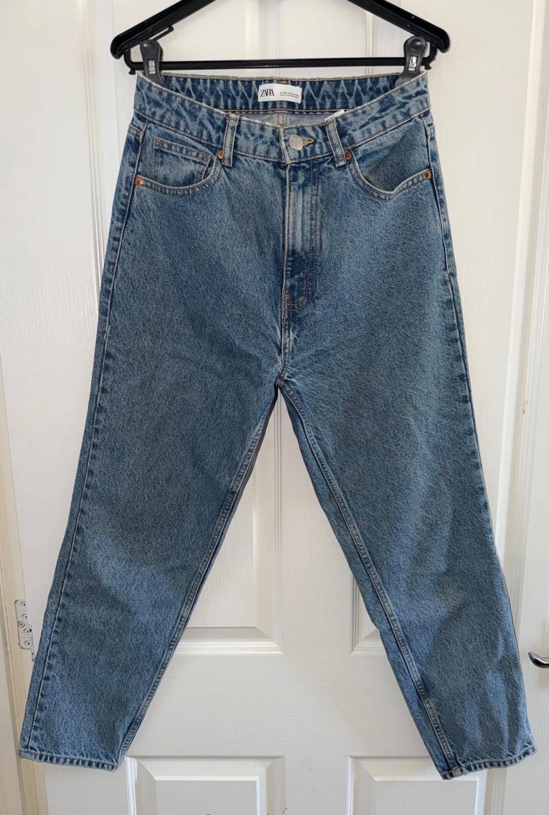 Blue jeans hanging on a black hanger in front of a white door. Label visible on the waistband.