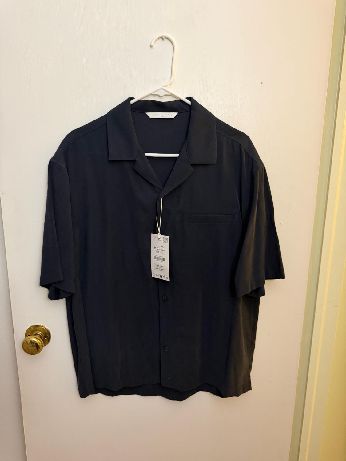 Dark, short-sleeved, button-down shirt with a pocket, hanging on a white hanger
on a white door with a gold doorknob.