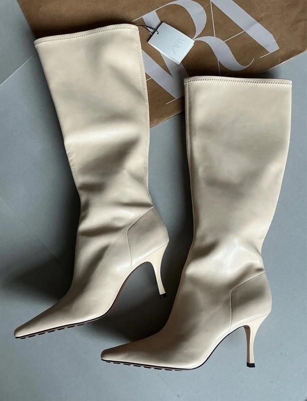 Beige high-heeled boots on a grey floor, with a white tag and a partially visible brown paper bag with white lettering.