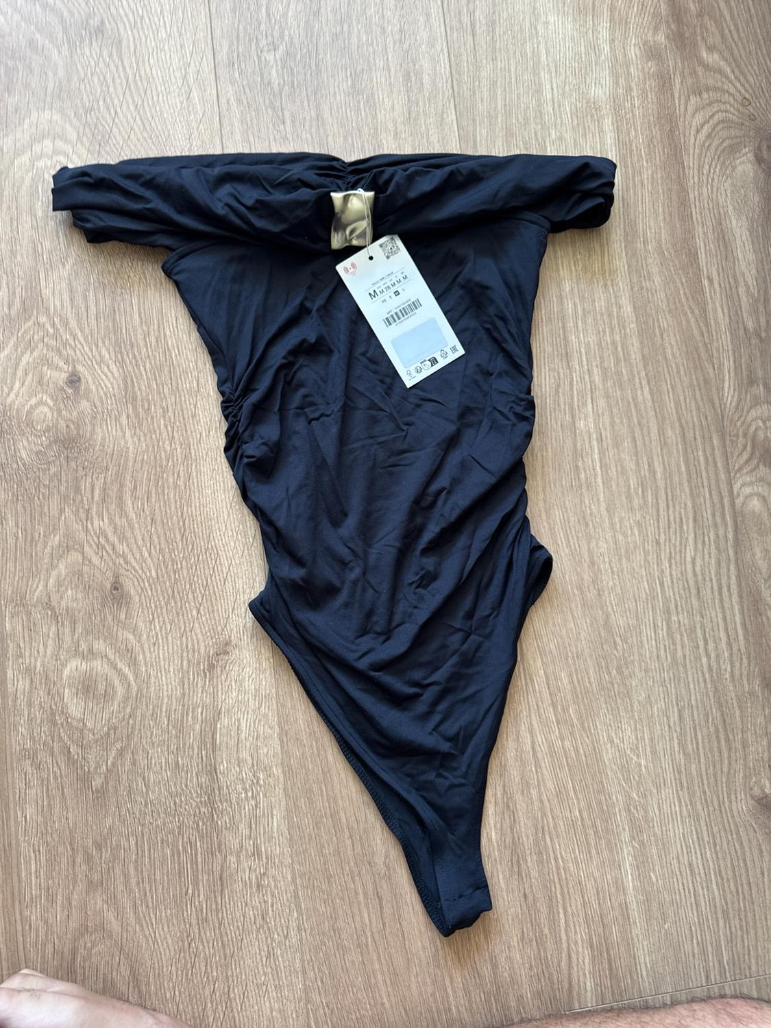 Black strapless ruched bodysuit with metallic detail on the neckline and white label, on a wooden floor.