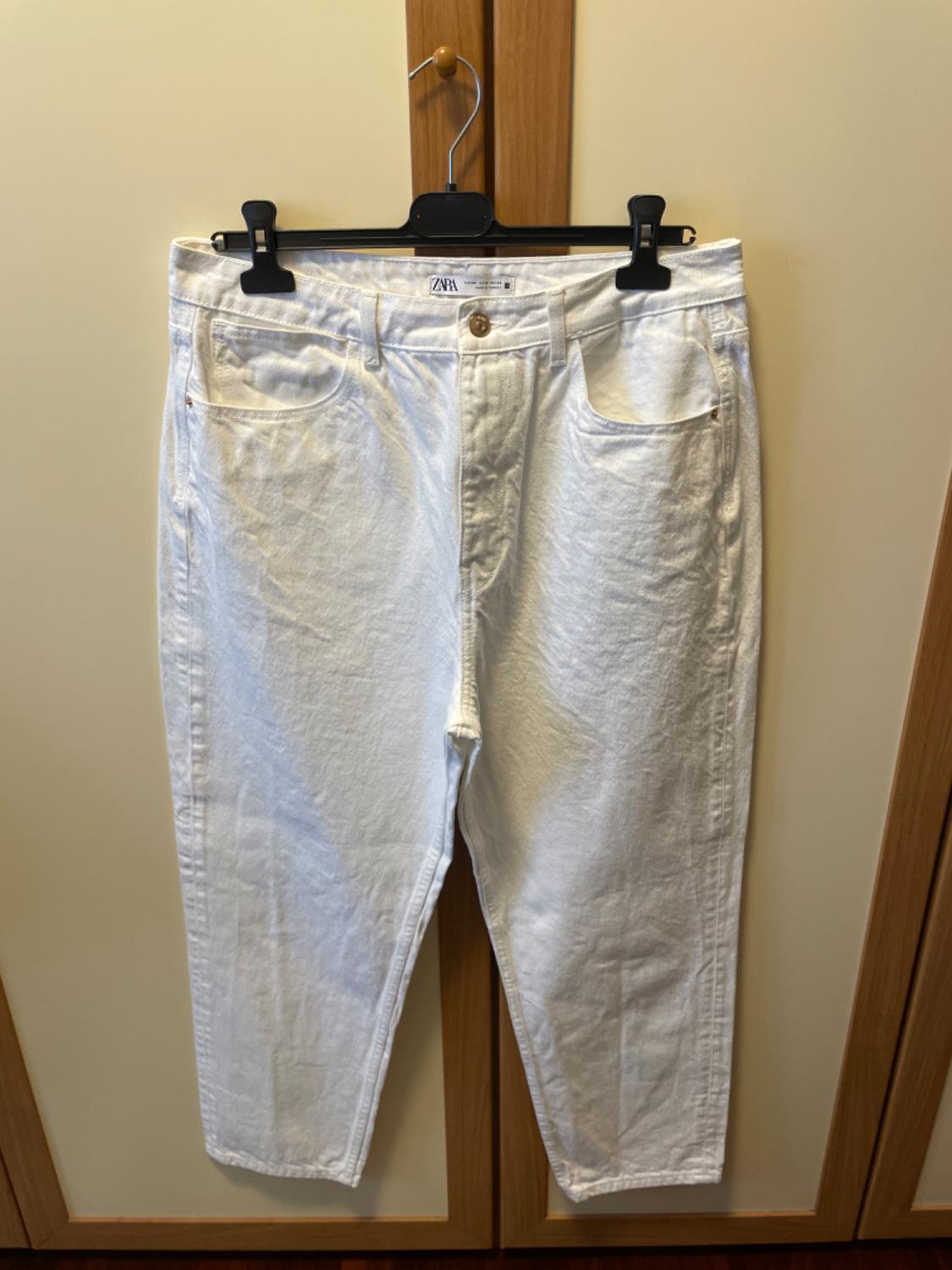 White trousers hanging on a black hanger in front of a light wood wardrobe. Visible label: "ZARA".