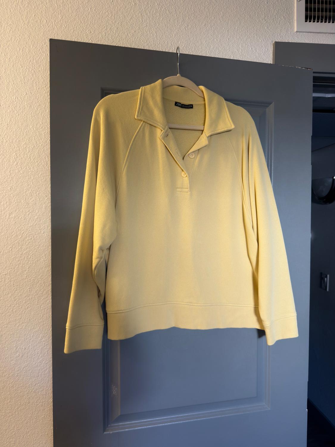 Yellow long-sleeved sweatshirt with a polo collar and buttons, hanging on a hanger over a grey door. Label with ZARA text.