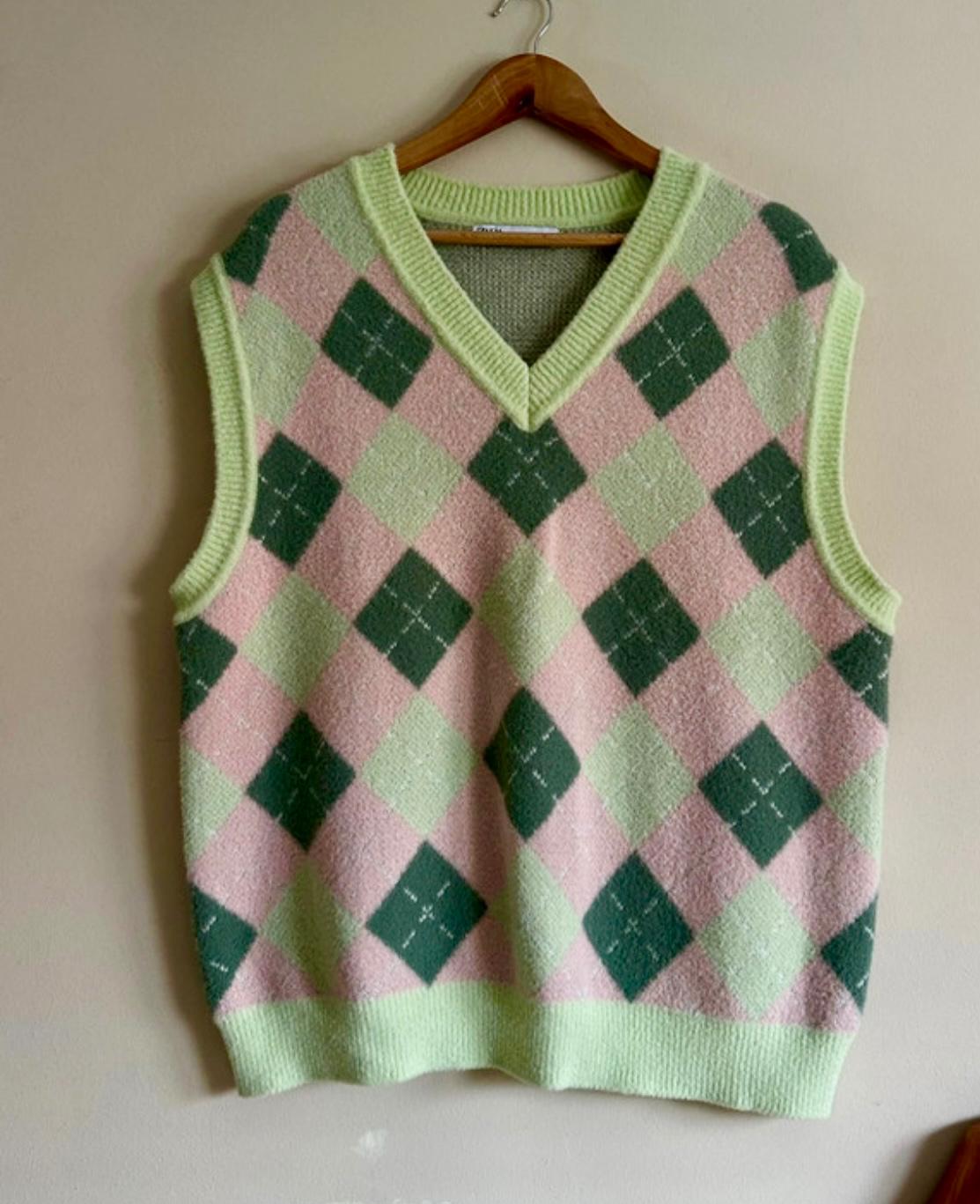 Knitted waistcoat hanging on a wooden hanger, with a argyle design in green and pink tones on a beige background.