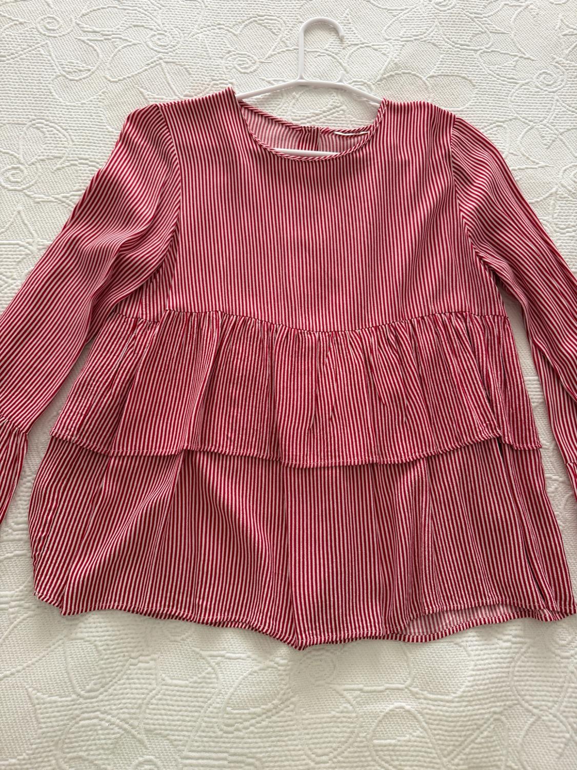 Red and white striped long-sleeved round-neck blouse with ruffles, on a hanger over a textured white bedspread.
