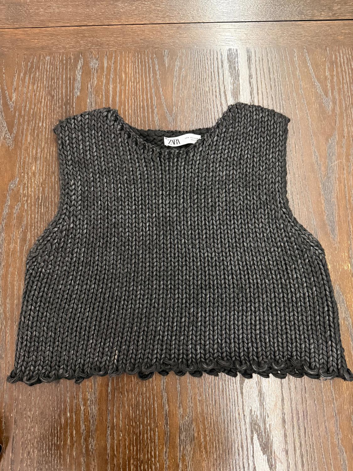 Black chunky knit vest on a wooden table. Visible tag with the text "ZARA".