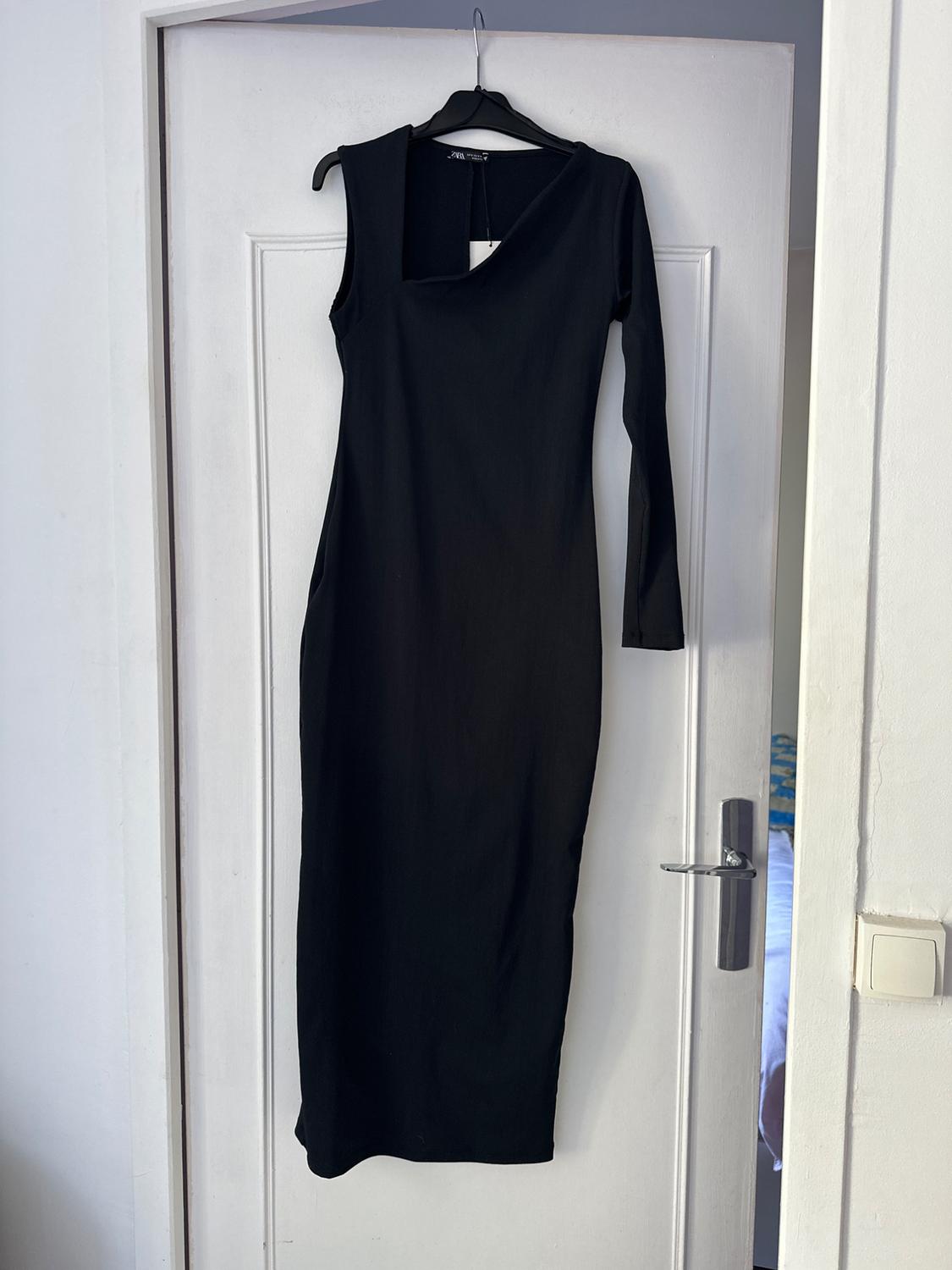Long-sleeved black dress hanging on a white door. Asymmetric design with one shoulder bare. Minimalist setting.